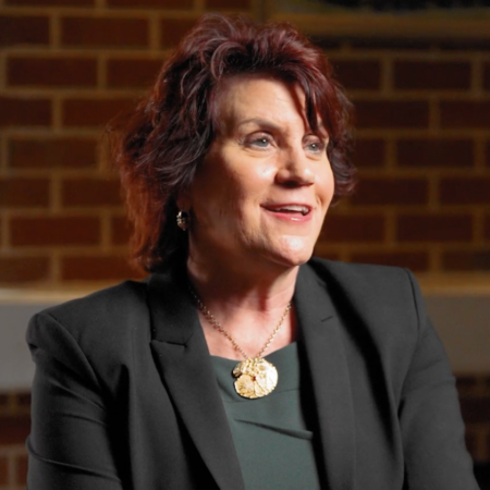 A woman with short dark hair wearing a black blazer and gold pendant necklace sits in front of a brick wall at the Center for Automotive and Mobility Studies, smiling and looking slightly to the side.